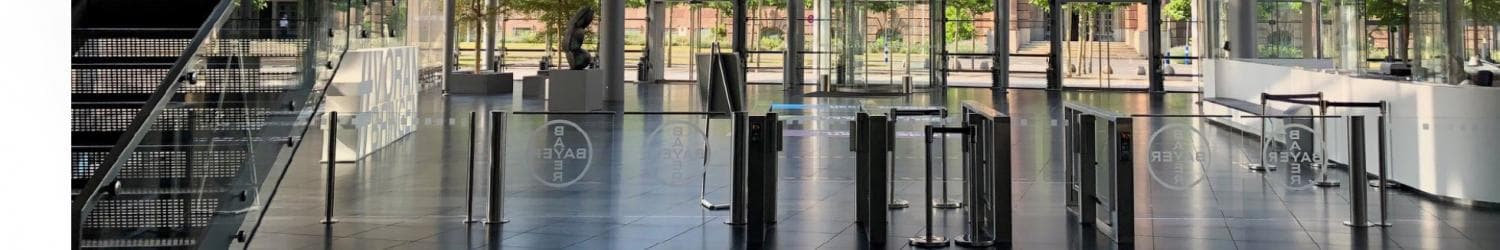 Modernes Atrium mit Glaswänden, Metallbarrieren, Treppen und Topfbäumen. Durch die Fenster ist ein großes historisches Gebäude sichtbar. Heller und geräumiger Innenraum.